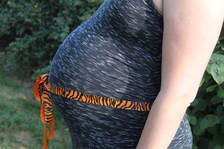 Profle of pregnant belly with orange tiger print ribbon tied around waist. Green bushes in background.