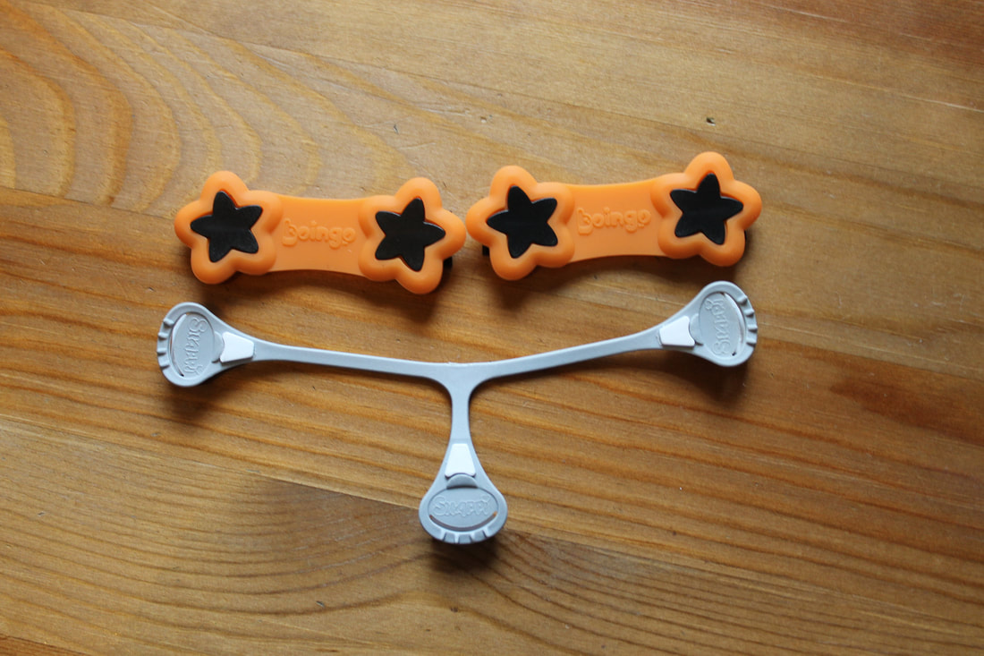 Diaper fasteners on wood background. Top fastener is orange Boingo. Consists of 2 bars with black stars at the ends of each bar. Bottom fastener is a grey Snappi. Snappi is T-shaped with a short bottom leg.